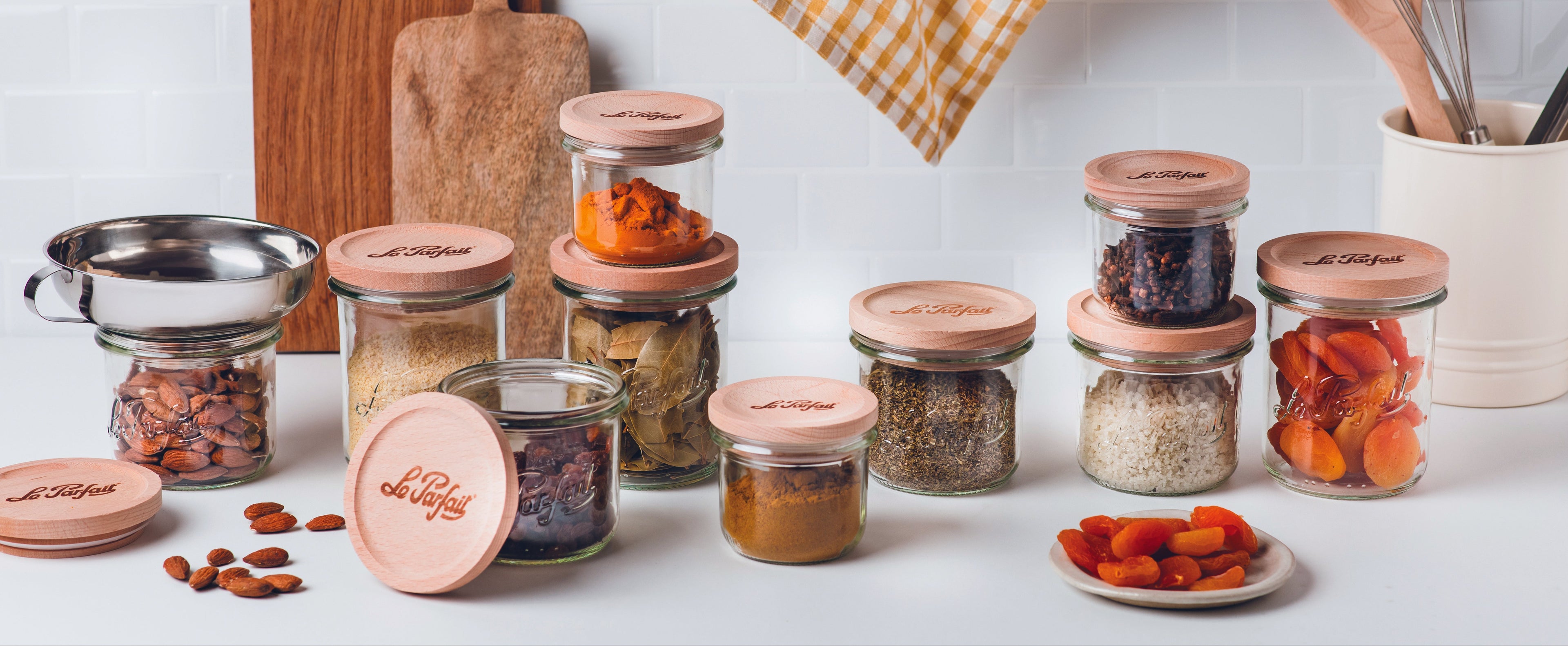 Collection of mixed sized glass jars with wooden lids on a kitchen counter, featuring food items.